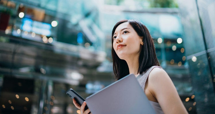 A woman holding a laptop and smartphone looks upward thoughtfully in a modern glass building with natural light and greenery in the background.